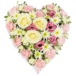 The photograph features a dense, heart-shaped floral tribute captured against a clear white background, its colours and textures standing out with gentle clarity, much like the arrangements created for services in Brent Cross and the surrounding neighbourhoods. The heart itself is fully covered with flowers, forming a rich, continuous surface of petals in soft pastel pink, creamy white, pale yellow, and touches of deeper magenta, all lightly accented by sprigs of green foliage. At the centre, several large cream-to-pale-yellow roses bloom in elegant spirals, their velvety petals layered in tight whorls that convey tenderness, respect, and enduring love. These roses rest in a generous bed of pastel pink carnations, whose ruffled petals overlap to create both the body and clear outline of the heart, giving it a cushioned, comforting appearance. Among the carnations, white spider chrysanthemums with slender, spiky petals and fresh green centres add lightness and intricate detail, like small bursts of brightness across the design. Deeper magenta-pink lisianthus blooms are dotted thoughtfully throughout, their softly fluted, bell-shaped flowers deepening the colour palette and bringing a quiet sense of refinement. Small, variegated green leaves and tiny buds peek through, reminding the viewer of the freshness and living beauty of the tribute. At the lowest point of the heart, a soft pink ribbon gently emerges, suggesting that this is a carefully prepared sympathy or funeral arrangement, intended to be placed beside a coffin, laid on a grave, or displayed next to cherished photographs. The overall impression is of a heartfelt, graceful floral heart, a fitting way for families in and around Brent Cross to express love, remembrance, and support in times of loss.