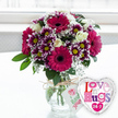 A charming love-themed gift set is captured on a pale, uncluttered surface, as if just placed down in a Brent Cross flat after a doorstep delivery. At the centre sits a rounded clear glass vase, its transparency revealing green stems gathered neatly in cool water. Around the neck, a piece of rustic, natural-coloured twine is tied into a small bow, supporting a slim white gift tag printed with a simple pink rose design and soft green script, giving the arrangement a personal, hand-prepared feel. Rising above the vase is a generous, rounded bouquet bursting with colour. Several large gerbera daisies in intense fuchsia and magenta dominate, their smooth petals radiating from dark, velvety centres that draw the gaze and add depth. Surrounding them, clusters of two-tone chrysanthemums bring lively contrast: rich purple petals fading to bright white at the tips and circling vibrant yellow centres like miniature suns. Creamy white roses are tucked in between, their petals still partly furled, lending a sense of gentle romance and elegance. Fine sprays of white baby's breath weave through the gaps, creating a soft, lacy texture that lightens the bolder tones. Here and there, fresh green foliage peeks out, framing the blooms and giving the bouquet roundness and volume. To the right of the vase, a heart-shaped foil balloon adds a playful note. Its border is shiny silver, catching the light, while the white centre is printed with mosaic-style lettering in pinks, purples and blues that spell a message of ongoing love and hugs. Behind the arrangement, the background is softly blurred and bright, suggesting daylight streaming through a nearby window, perhaps overlooking Brent Cross Shopping Centre or the streets towards Cricklewood. The overall effect is joyful and affectionate, perfect for birthdays, romantic surprises or simply letting someone know they are loved, with flowers delivered locally across Brent Cross.