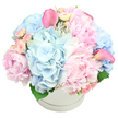 A round white hatbox, immaculate and understated, is filled to bursting with a lush arrangement of pastel flowers, set against a plain white background that highlights every petal. Dominating the bouquet are large, soft pink peonies, some fully open and others in mid-bloom, their ruffled, layered petals creating a plush, cushiony effect that exudes romance and warmth. Interspersed among them are full heads of light blue hydrangeas, composed of myriad tiny florets that gather into cloudlike domes, lending a cool, tranquil counterpoint to the pinks and evoking the calm of an early morning around Brent Cross before the shops open. Nestled between the larger blooms are blush spray roses, still tightly furled, their small buds adding fine detail and suggesting beauty yet to unfold. Two vivid pink calla lilies extend upward from the arrangement, one reaching elegantly toward the left, the other toward the right, their sleek, trumpet-shaped flowers introducing height and a modern, sculptural element that would suit contemporary interiors in the new developments around the North Circular. Discreet glimpses of dark green foliage are visible at the base, offering subtle contrast and grounding the pastel palette. The flowers are densely packed, creating a rounded, abundant mound that appears to overflow slightly from the hatbox, which features a gentle ridge circling its upper edge. The overall feel is one of tender sophistication and gentle celebration, well-suited for welcoming a new baby, sending congratulations, or offering a comforting, elegant gesture to friends and family in Brent Cross and neighbouring parts of North West London. It looks like a bouquet designed to bring a moment of peace and beauty into everyday life.