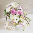 A refined wedding-style centerpiece in shades of blush pink and pure white brims from a round white wicker basket, evoking the atmosphere of an elegant Brent Cross celebration. At its centre, large pastel pink roses unfurl in layered spirals, each velvety petal curving softly outward, creating a sense of fullness and romantic warmth. Tucked among them, white Phalaenopsis orchids open their broad, curved petals, framing golden yellow and delicately speckled centres that add a hint of tropical luxury. Between the roses and orchids, ruffled white Lisianthus blossoms introduce subtle texture and depth, some featuring a gentle wash of lavender-purple along their edges, giving a cool undertone that balances the blush tones. Slender stems of pale pink stock rise and then cascade slightly over the lip of the basket, their clusters of tiny blossoms producing feathery plumes that add height, light fragrance, and effortless movement, as if caught in a soft breeze crossing nearby Brent Cross Park. Wisps of baby's breath appear like a fine veil throughout the design, thousands of tiny white blooms softening the spaces between larger flowers and lending a dreamy, cloudlike quality. Fresh, glossy green leaves peek through at intervals, with a few delicate buds and stems arching over the edge, adding a natural, slightly cascading finish. The tightly woven white basket sits atop a clean, bright white surface that could be a dressed wedding table or a modern North London dining room, accompanied by two decorative woven spheres in pale wood tones whose open, lattice structure echoes the basket's texture and adds sculptural interest. Behind, a neutral beige, linen-textured backdrop creates a warm, understated setting that throws the flowers into gentle relief. The overall mood is romantic, calm, and celebratory, ideal as a centerpiece for weddings, vow renewals, or special gatherings around Brent Cross.
