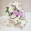 An exquisitely balanced arrangement of wedding-style flowers is presented on a pristine white surface, evoking the soft elegance of a quiet evening gathering near Brent Cross. Dominating the display are several stems of Phalaenopsis orchids, their large white petals arching outward like wings caught mid-flight. The creamy surfaces of the petals are contrasted by the orchids' golden-yellow and orange centres, which glow warmly at the heart of each bloom and draw the viewer into the design. Surrounding them, full-headed lavender-pink roses open in perfect spirals, each petal velvety and carefully layered, creating a sense of classic romance and time-honoured beauty. Interspersed among the larger flowers, sprays of delicate white baby's breath add lightness and a fine, lace-like texture, softening the transitions and filling small spaces with airy detail. Elegant stems of light purple lisianthus, some fringed in deeper violet, rise gently between the roses and orchids, adding ruffled contours and a subtle variation of tone. From the lower edges of the bouquet, clusters of pastel lavender-pink blossoms, reminiscent of stock or sweet peas, drift downward, giving the composition a graceful, cascading movement. Fresh green foliage, both broad and narrow, provides structure and a vivid counterpoint to the soft pastels, grounding the arrangement visually. All of this is gathered into an innovative white vase constructed from slender, interwoven strips arranged in a spherical lattice, allowing glimpses of the stems within and lending a contemporary sculptural quality. Two matching woven orbs rest beside the vase, echoing its design and reinforcing the modern aesthetic. The softly textured beige background and diffused lighting emphasise the gentle interplay of colour and form, creating an atmosphere of serenity and refined celebration. It is easy to imagine this piece adorning a wedding reception in a Brent Cross hotel or arriving as a same-day anniversary surprise from Brent Cross Flowers, carrying a message of enduring love and thoughtful elegance.