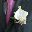 A close-up photograph shows a sharply tailored charcoal or near-black suit jacket, the left lapel turned slightly toward the camera so every detail of the formal attire is visible. A pale grey dress shirt with a fine, almost woven texture peeks through, its soft pattern adding quiet interest beneath a rich magenta-to-plum tie, perfectly knotted and resting smoothly against the chest. Pinned precisely to the lapel is a striking boutonniere built around a flawless white Phalaenopsis orchid, its wide, velvety petals gently curved and glowing against the dark fabric. At the flower's heart, a warm yellow and soft orange centre adds a tiny burst of colour, like a pool of light. Around the orchid, clusters of tiny pale lilac blossoms and airy white baby's breath create a cloud of delicate texture, supported by a few polished deep green leaves that frame the design. Just above the open bloom sits a single light green orchid bud, still closed, suggesting new beginnings and the quiet anticipation of a wedding morning somewhere near Brent Cross or along the Finchley Road. A slim lavender or mauve satin ribbon is tied into a small, neat bow at the base, securing the stems just above the edge of the suit's breast pocket. The lighting is gentle and even, emphasising the satin sheen of the tie, the smoothness of the petals, and the crisp lines of the suit. Overall, the image conveys understated elegance and careful coordination, as if prepared by an experienced local florist for a groom about to walk into a ceremony in one of Brent Cross's nearby venues, full of calm confidence and romantic expectation.