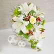 A graceful floral centerpiece rests on a smooth white tabletop, its refined style perfectly suited to a modern wedding reception or engagement dinner in Brent Cross. The flowers emerge from a round white woven basket, the wicker forming a soft lattice that cradles the composition and adds gentle texture. At the core, large white lilies open in full trumpet shapes, their petals silky and luminous, touched with faint speckling that draws attention to the central yellow anthers dusted with pollen. Slightly above them, a single cream-yellow rose, still somewhat budded, stands as a subtle focal point, its layered petals promising to unfurl further. Drift­ing from the right-hand side, a cascade of white phalaenopsis orchids trails elegantly downward, each bloom perfectly poised, creating a flowing, waterfall-like line that suggests movement and elongates the design. Around these feature blooms, an abundance of green foliage and textural accents fills the basket: feathery sprigs, slender grass-like leaves, and bright lime-green pom-pom flowers that add a plush, contemporary touch. Threaded delicately between the larger flowers, tiny white blossoms reminiscent of baby's breath create a fine, cloud-like veil, while clusters of rich red berries offer vibrant pops of colour, similar to festive arrangements delivered around Brent Cross at Christmas and special occasions. To the left of the main arrangement, two decorative white spheres sit on the table, one larger and one smaller, fashioned from thin, interwoven strips that form open, airy patterns and echo the basket's craftsmanship. The soft beige background, textured like fabric or linen, enhances the sense of depth while keeping the focus on the flowers, producing an overall feeling of purity, serenity, and joyful celebration.
