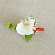 A meticulously crafted wedding boutonniere is laid carefully on a softly textured light beige linen-style cloth, the neutral background allowing every detail of the flowers to stand out. At the heart of the design sits a single white phalaenopsis orchid, its broad, velvety petals opening in a calm, sculptural curve. The orchid's throat glows with soft yellow and hints of warm reddish-orange markings, giving depth and character to the otherwise pure white bloom. Around it, tiny white baby's breath flowers gather like a delicate cloud, their miniature blossoms adding a gentle, lace-like texture. To the left, two round lime-green chrysanthemum buds, tightly packed and slightly spiky, provide a fresh, modern contrast, their crisp green tones echoing the lively feel of spring mornings around Brent Cross and the nearby parks. A broad, deep green leaf forms a natural base beneath the orchid, while a smaller darker leaf peeks out close to the stems, grounding the composition with rich foliage. The stems are neatly bound with a vivid crimson red satin ribbon, wrapped smoothly and finished with a small, tidy bow that introduces a bold ribbon of colour and a sense of celebration. Soft, even lighting enhances the natural sheen of the orchid and the subtle weave of the fabric, making this boutonniere feel quietly luxurious yet approachable. The piece evokes the mood of a joyful ceremony, ideal for a groom or groomsman heading to a Brent Cross registry office wedding or a nearby hotel celebration, carrying a discreet but unforgettable touch of floral elegance on their lapel.