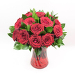 A luxurious display of deep crimson red roses fills the frame, each bloom clustered closely with a sense of abundance and intention, as though gathered by a careful florist in Brent Cross for a truly special moment. The roses are at different stages of opening: some remain tightly furled at the centre, while others unfurl in velvety spirals, revealing layers of rich, ruby-toned petals that almost seem to glow against the crisp white background. Lush, dark green foliage, likely Ruscus or similar glossy greenery, is woven between the blooms, adding depth and a cool, polished contrast that makes the red appear even more intense. The bouquet is cradled in a translucent ruby-red glass vase with a subtle twisted or spiralled pattern etched into its surface, catching and bending the light so that reflections dance across the glass. Through the coloured glass, the rose stems can be seen, slightly softened and tinted by the red hue, giving the whole arrangement a cohesive, jewel-like quality. The vase flares gently at the top and rests on a solid base, grounding the otherwise soft, romantic composition. Bright, even lighting highlights the freshness of each petal and leaf, as if the bouquet has just been arranged for same-day delivery to a home near the shopping centre or a cosy flat off Hendon Way. The overall impression is one of classic romance and refined celebration, perfect for anniversaries, Valentine's gestures, or any heartfelt declaration of love in and around Brent Cross.