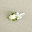 This boutonnière showcases a single, beautifully formed ivory rose lying gently on a pale, textured cloth, captured in soft, even light that highlights every curve. The rose is at the perfect stage of opening, its petals beginning to unfurl from a tight centre into a layered spiral, each one thick and velvety, with a creamy tone that feels warm and inviting. Encircling the rose, stems of gypsophila - baby's breath - sprawl delicately, each thread-like stem tipped with clusters of tiny, white flowers that resemble miniature clouds. These airy blooms add softness, suggesting the kind of gentle romance that often colours wedding days around Brent Cross and the neighbouring streets of Golders Green. Fine, glossy blades of ornamental grass loop gracefully around the rose and gypsophila, their vivid green hue providing contrast and a hint of natural spontaneity, as though the boutonnière were freshly gathered. Beneath this arrangement, the stems are neatly enclosed in a sheath of white satin ribbon, wrapped tightly with steady, practiced hands. A small, tidy bow sits near the base, its smooth loops resting flat and secure, ready to be pinned to a lapel. The background fabric, subtle in texture and neutral in colour, ensures the flowers remain the focal point, every petal and flower head standing out clearly. The overall feel is classic, calm, and deeply personal, ideal for a groom, usher, or honoured guest at a ceremony near Brent Cross or along the Northern line corridor. Imagining the faintly sweet, green scent of the rose and foliage, one can almost sense the quiet excitement of dressing for a long-anticipated celebration.