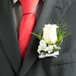 In this close-up, an elegant boutonniere is pinned neatly to the lapel of a charcoal grey suit, capturing the spirit of a classic Brent Cross wedding or prom. A single white rose forms the heart of the design, its petals creamy and luminous, curling outward in a natural spiral that feels both tender and confident. The bloom's surface appears velvety and smooth, with subtle shadows deepening in the folds, like delicate brushstrokes of soft grey. Gathering around its base, fine sprays of white baby's breath add a cloudlike edge, their tiny, star-shaped blossoms offering a gentle contrast in both size and texture. The baby's breath softens the transition from the structured suit to the organic curves of the rose, suggesting a meeting point between city refinement and the quieter greenery found near Brent Cross Park and the banks of the nearby River Brent. Providing further depth, slender fern fronds and curved strands of glossy green foliage arc gracefully along the lapel, giving the arrangement a sense of flow as if it were gently stirred by an indoor breeze at a local reception venue. A slim white satin ribbon wraps the stems and finishes in a small, precise bow, the fabric's subtle sheen echoing the polished shoes and cufflinks often worn with such a suit. The mood is one of poised anticipation, as though the wearer is moments from walking into a ceremony at a nearby hotel or family gathering in Hendon or Golders Green. The boutonniere feels personal and intentional, a modest yet meaningful flourish that conveys thoughtfulness, tradition, and a quiet, heartfelt joy on a special North London day.