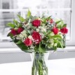 A lavish bouquet of red roses, ruffled red carnations, and crisp white alstroemeria stands proudly in a clear glass vase on a bright white tabletop, bathed in soft daylight from a large window. Each rose, some fully open with velvety petals and others still in bud, feels like a promise of affection, while the carnations add generous volume and a traditional, comforting charm. Between the bold red blooms, clusters of white alstroemeria, many still in tight buds with pale green tips, create a gentle rhythm of anticipation as they wait to unfurl. Tiny white sprays of gypsophila, or baby's breath, drift lightly through the arrangement like a delicate mist, softening the overall look and catching little glints of light. The bouquet is framed by lush, layered greenery: broad, glossy dark green leaves form a structured collar around the base, while more slender, lighter foliage threads upward, adding texture, depth, and a feeling of fresh cut stems straight from a local Brent Cross market stall. Through the clear vase, the neatly cut stems are visible, standing in clean water that reinforces the impression of just-arrived freshness, ready for same-day delivery across Brent Cross and the nearby streets towards Hendon and Golders Green. Behind the arrangement, the window view is softly blurred and almost monochrome, so the vivid reds, whites, and greens seem to glow against the neutral background. This composition feels like a heartfelt message made visible-perfect for an anniversary dinner at home, a romantic gesture after a walk through Brent Cross shopping centre, or simply to brighten a quiet living room with the joy of fresh flowers and the reassuring presence of thoughtfully crafted design.
