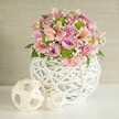 Awash in gentle natural light, a handcrafted floral centerpiece brightens a Brent Cross event table, exuding warmth, modern style, and the soft romance of spring celebrations. The generously rounded bouquet blends pastel pink and lavender roses, each bloom layered and plush, calling to mind a stroll through Brent's community gardens after rain. Scattered within are pink alstroemeria, easily distinguished by their delicate flute shapes and decorative inner streaks, adding elegance and lively detail. Vivid lime green button chrysanthemums emerge throughout, their compact forms providing cheerful contrast, while billowy white baby's breath imparts an airy, almost whimsical quality. All are set against a foundation of dark emerald leaves-textured, glossy, and grounding. The arrangement rests in a unique white vase, its open-weave structure skillfully intertwining slender strips in an artful, modern pattern, suggesting craftsmanship found in the heart of Brent Cross. Completing the display, two off-white, woven decorative spheres-one large, one small-are placed alongside, their airy constructions and natural tones mirroring the vase's intricate latticework. A softly textured beige wall forms the backdrop, letting the vibrant florals and creative textures take center stage. This setting resonates with quiet celebration and heartfelt care, making it a perfect choice for local Brent Cross weddings, anniversary feasts, or as a centerpiece that reflects the vibrant, diverse spirit of the community.