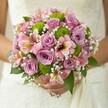 Basking in soft daylight, this exquisite bridal bouquet-crafted for a Brent Cross celebration-embodies the spirit of new beginnings. Plush roses in pastel pink and whispery lavender spiral open at the heart, exuding a sweet, heady fragrance that mingles with the springlike freshness of nearby Welsh Harp Reservoir. Accents of lime green button chrysanthemums provide a lively lift, their rounded forms and vibrant hue adding energy and contemporary style to the timeless charm of the roses. Scattered amongst the central blooms, pink alstroemeria lilies reveal delicate trumpet-shaped petals with distinctive streaks of yellow and berry, suggesting both delicacy and playfulness. Silky tufts of white baby's breath drift through the bouquet, creating airy lightness akin to gentle clouds rolling above Brent Cross's green spaces. Deep green leaves peek from the borders, providing definition and natural structure, while the entire creation is cocooned in the bride's gentle hold-her hands, adorned with a simple ring, securing the wrapped stems just above the flowing folds of a lace gown. This bouquet is a whisper of romance, expertly composed for joyful, heartfelt gatherings in Brent Cross and the wider North West London community, radiating warmth, love, and timeless beauty.