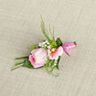This boutonniere appears like a tender message of affection, crafted with the same attention to detail you might expect from a bespoke florist serving the Brent Cross community. At the centre sits a cool pink rose, its layers of velvety petals unfolding in a gentle spiral, with the outer edges curling back just enough to suggest openness without losing their refined form. The colour deepens from soft blush at the core to a more confident pink along the rim of each petal, the surface catching the light and creating subtle highlights and shadows. Supporting the rose are pastel alstroemeria blooms, their petals narrow and slightly flared, featuring delicate markings and warm yellow centres that bring a touch of sunshine to the arrangement. Fine white gypsophila is sprinkled throughout, each tiny blossom like a soft star, adding a dreamy, cloudlike quality. Slender, fresh green fronds and leaves extend around the flowers, bringing structure and a crisp, garden-fresh texture, as though the stems were snipped from a nearby garden off Hendon Way or near Brent Cross station. Everything is held flawlessly together by a neatly wrapped pink ribbon, smooth and glossy, which encases the stems in a streamlined finish suited to a tailored suit lapel. Resting on a softly woven background, the flowers stand out clearly, bathed in gentle light that enhances their romantic hues. Imagining this boutonniere pinned to a jacket at a wedding reception in a local hotel or a prom gathering just off the North Circular, you can sense its intention: to convey thoughtfulness, tenderness, and pride in a way that feels personal and sincere. It is a small floral accent that elevates a moment, lending the wearer an air of quiet elegance and heartfelt celebration.