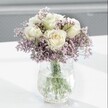 A close-up photograph shows a serene white and lavender bouquet glowing softly against a pale, blurred background, as if placed on a bright windowsill in a quiet Brent Cross home. At the heart of the design are several creamy white roses, their petals layered and gently unfurling, with the faintest suggestion of pale buttery yellow deep in the centres that hints at absolute freshness. Each rose head is full and velvety, the kind of bloom that a skilled florist selects carefully at just the right stage so that it will open beautifully over a few days. Around them, like a mist, spreads an abundant halo of tiny lavender and lilac-hued statice flowers. Their papery, cloud-like clusters create a soft, romantic haze, adding both fullness and a delicate, almost featherweight texture that contrasts with the smooth, voluptuous rose petals.

The bouquet is gathered into a clear glass vase with a distinctive crackled or ice-like texture, the surface catching the light in small, sparkling lines that feel almost frosted. Through the water inside, the straight green stems are clearly visible, cleanly cut and neatly arranged, suggesting professional preparation and very recent conditioning. Everything rests on a smooth, white tabletop that reflects a little of the daylight and throws a gentle shadow beneath the vase, reinforcing the impression of calm and balance.

The overall mood is peaceful and understated, like a quiet morning walk through the greenery near Brent Cross or along local residential streets before the city fully wakes. This arrangement feels ideal for a thoughtful sympathy gesture, a gentle get-well wish, or to bring a sense of calm elegance to a bedroom or study, its soft colours and relaxed shape offering comfort without ever feeling overpowering.