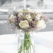A full, rounded bouquet of creamy white roses and lilac statice stands in the centre of the image, captured as if resting on a bright kitchen table in a Brent Cross flat. Around a dozen white roses form the heart of the design, each bloom at that perfect stage where the petals are just beginning to unfurl, revealing layered spirals of soft ivory and warm cream. Their velvety texture looks almost touchable, with edges that catch the daylight streaming in from an unseen window. Threaded between the roses, clouds of delicate, light lavender statice create a gauzy halo, each tiny papery flower adding a fine, feathery texture that softens the more structured rose heads. The contrast of the plush, domed roses and the airy sprigs makes the bouquet feel both elegant and gentle, ideal for moments that need quiet reassurance or refined celebration. All the stems are gathered neatly in a tall, clear cylindrical glass vase, the water so clean it almost disappears, leaving the fresh green stems to provide a cool vertical rhythm. The vase is placed on a smooth, white surface that reflects subtle shadows and faint glimmers of light, adding to the sense of calm. The background is softly blurred, suggesting an airy indoor space-perhaps a living room near Brent Cross Shopping Centre or a calm office off the North Circular-bathed in natural light. The overall scene conveys peaceful sophistication, perfectly suited to sympathy flowers, a thoughtful anniversary surprise, or a serene thank-you gesture delivered locally around Brent Cross.