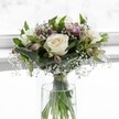 An elegant white rose bouquet stands in a simple cylindrical glass vase, bathed in soft daylight that feels like it could be streaming through a bright Brent Cross apartment window. Three fully opened, creamy white roses form the heart of the arrangement, their petals gently unfurling in layered spirals, with subtle ivory centers that suggest perfect freshness and quiet purity. Around these focal blooms, rich emerald foliage curves and folds, some leaves broad and structured as if carefully cradling the flowers, others more delicate and feathery, adding a sense of depth and natural movement. Threaded between the greenery, tiny white gypsophila - classic baby's breath - drifts like light mist, its fine stems and miniature blossoms creating a cloud-like halo that softens the whole design. Nestled discreetly among the roses are alstroemeria buds and blossoms in muted purple and pale pink tones, some still closed, others just beginning to open, giving hints of colour that promise more beauty to come without overwhelming the calm, white palette. The stems, neatly cut and gathered, are visible through the clear water of the vase, appearing to be lightly tied with a slim ribbon that holds them in a tidy column. The background is softly blurred and almost entirely white, suggesting a luminous window or bright conservatory, and this gentle glow highlights every petal and leaf texture. The overall mood is peaceful and refined, a compact, rounded bouquet that would feel at home bringing comfort to a quiet living room in Brent Cross, resting on a bedside table after a thoughtful delivery, or marking a moment of remembrance with soft grace and understated elegance.