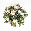 In this overhead image, a thoughtfully arranged sympathy bouquet sits centered on a pure white surface, its circular shape and restrained palette conveying composure and calm. The focal blooms are creamy white roses, their petals smooth and delicately curved, unfolding in symmetrical spirals that draw the eye in gentle stages toward the heart of each flower. Some roses are open and expansive, revealing soft shades of ivory and a hint of peach deep inside, while others remain more tightly gathered, adding a sense of balance and quiet potential. Threaded among the roses, purple alstroemeria buds and just-opening flowers bring subtle colour and texture. Their petals show gradients of lavender, lilac, and green, with some early signs of the fine streaks characteristic of this variety, as though the bouquet is caught between stillness and gradual bloom. Throughout the design, small plumes of white baby's breath weave in and out, their countless tiny blossoms creating a gossamer veil that softens every edge and gap. Dark green foliage forms a cradle beneath and around the flowers, its broad, smooth leaves slightly glossy in the light, holding the bouquet in a rounded, cohesive form. The overall impression is one of quiet dignity, like an arrangement that might rest on a low table in a Brent Cross flat or beside a framed photograph in a family home. It feels fresh and carefully composed, without showiness-simply an honest, beautiful cluster of blooms ready to offer comfort. The soothing mix of whites, gentle purples, and rich greens allows this bouquet to speak softly of remembrance, support, and the wish for peace in a difficult moment.