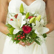 This bridal bouquet, photographed against the detailed lace of a wedding dress, feels like a moment of stillness on a busy Brent Cross morning, just before guests arrive and music begins. At its heart, a luxurious red rose blooms in perfect symmetry, the petals velvety and dense, drawing the eye to their deep, romantic hue. Around this focal point, white lilies spread their star-like petals with quiet grandeur, their creamy surfaces reflecting the light in soft, subtle highlights. Some lilies are wide open, showing delicate stamens dusted with pollen, while a few buds remain closed, suggesting the unfolding of the day ahead. Feathery sprigs of gypsophila weave through the arrangement, their tiny blossoms forming a soft mist between the bold flowers, lending a classic, almost timeless bridal feel. Bright red berries punctuate the greens with cheerful dots of colour, adding a playful, seasonal edge that brings to mind winter weddings or celebrations near Brent Cross's tree-lined streets. Mossy green textures and dianthus tufts nestle near the base of the blooms, giving a touch of garden freshness and tactile richness, as if the bouquet has been gathered from a well-tended local park. Slender grasses and light, arching foliage create a gentle halo around the bouquet, lightening the silhouette and making it appear slightly airy and free. Tied neatly at the stems, the bouquet looks comfortable and balanced in the bride's hands, designed with both beauty and practicality in mind. Altogether, the combination of creamy lilies, passionate red rose, delicate filler flowers, and textured greens conveys romance, joy, and the cherished intimacy of a wedding day.