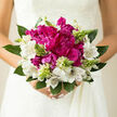 In this image, a carefully crafted bouquet rests between gently clasped hands, its colours and textures capturing the romantic spirit of a Brent Cross wedding. The focal point is a cluster of sumptuous magenta peonies, their petals plush and plentiful, overlapping in dense, soft layers that invite the fingertips to trace their curves. The tone of magenta is rich yet sophisticated - more like deep raspberry than bright fuchsia - giving the bouquet an elegant, timeless heart. Surrounding these central blooms, white alstroemeria form a delicate frame. Each flower is trumpet-shaped, opening into a soft star, and is adorned with fine burgundy streaks and tiny dashes, like ink strokes across a canvas, adding subtle depth and detail.   

Between peonies and alstroemeria, fine stems topped with pale green buds emerge. Some buds are tightly closed, others beginning to loosen, suggesting the gentle unfolding of a new season, much like the changing days seen from the bridges and streets around Brent Cross and Cricklewood. The foliage encircling the bouquet is lush and deep green, the leaves smooth, slightly glossy, and gently arched, providing structure and a natural frame for the brighter blooms. Soft, natural light, reminiscent of a clear afternoon over the North Circular, bathes the bouquet, catching the edges of each petal and leaf, and creating gentle contrasts rather than harsh shadows. The effect is one of calm elegance, as if time has slowed in the moment before a bride steps into the aisle. Though the fragrance remains unseen, it is easy to imagine the delicate mix of cool greenery and mild floral sweetness rising from the bouquet. This arrangement would feel equally at home in an intimate ceremony near Brent Cross or a quiet celebration at home in NW11, carrying with it a sense of poised joy and heartfelt intention.
