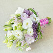 A refined hand-tied bouquet is displayed on a lightly textured beige fabric, angled so the stems point toward the lower corner, as if just set down by a bride pausing for a moment of calm. The composition is a serene blend of white, lavender, and deep purple with fresh greens, creating a look that feels both polished and natural, like a stroll through a landscaped garden near Brent Cross. Dominating the arrangement are lush white lisianthus flowers, some fully open to show creamy, almost buttery yellow centres, others still unfurling from pale green buds. Their soft, ruffled petals stack delicately on one another, forming cloud-like blooms. Interlaced among them are small clusters of pastel lavender-pink blossoms and airy, lilac-toned phlox that bring a veil of soft colour and lightness. Eye-catching accents of starburst purple alliums introduce a striking architectural element: each globe is made of tiny pointed florets, giving the bouquet an artistic edge. Verdant greenery, including long, tapered leaves, curled strands of bear grass, and lighter green spiky textures, weaves in and out, adding rhythm, depth, and a sense of motion. The stems are neatly trimmed and tied, then wrapped in a bold magenta-fuchsia satin ribbon that gleams against the greens and whites. Along this handle, a line or delicate garland of white pearls adds a touch of luxury reminiscent of bridal jewellery. The overall impression is of modern romance and quiet sophistication, ideal for a wedding held around North Circular Road venues or for same-day delivery in Brent Cross to mark anniversaries, engagements, or any intimate celebration of love.