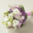 On a neutral, softly textured tabletop reminiscent of Brent Cross's understated elegance, this lush, hand-tied bouquet radiates romance. Prominently featured are sumptuous white Lisianthus blossoms-petals unfurling in luxurious layers, the centres kissed by soft gold-paired with unopened ivory buds for freshness that echoes the calm green spaces near Hendon Park. Galea-like pastel pink and lavender Stock cluster throughout, their ruffled forms adding depth and a hint of whimsy, while vivid, spherical Allium 'Star of Persia' in deep purple add modern flair and sculptural intrigue. Emerald green, arching foliage interlaces between the blooms, creating a movement and vibrancy that mirror Brent Cross's blend of city and serenity. The stems are artfully wrapped in pastel fuchsia ribbon banded with delicate pearls, catching the light and imparting a sense of celebration. Every element-from the smooth Lisianthus petals to the starry Allium crowns-comes together in a classic, rounded composition, ideal as a bridal bouquet or centrepiece for special events. This arrangement captures the spirit of Brent Cross-fresh, refined, and full of subtle joy.
