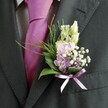 A close-up wedding portrait focuses tightly on the upper body of a man in a sharply tailored charcoal suit, his lapel adorned with a carefully crafted buttonhole that brings soft colour and romance to the dark fabric. The suit jacket, in deep grey verging on black, shows a fine woven texture along the lapel and sleeve, while a pale dress shirt with a discreet grid pattern peeks through beneath a bold magenta silk tie, its knot smooth and lustrous in the gentle light. Pinned neatly to the left lapel is a delicate boutonniere composed of slender, unopened creamy buds that could be snapdragons, rising above a cluster of pastel pink and lavender flowers reminiscent of sweet peas or stocks. Tiny white stars of baby's breath are threaded throughout, adding misty lightness, with a few deeper purple florets deepening the palette. At the base, glossy dark green leaves provide structure, offset by wisps of brighter, feathery foliage that arc outward like soft fronds of asparagus fern, giving the piece movement and air. A small lavender satin ribbon is tied in a tidy bow around the stems, echoing the lilac tones in the petals and sitting just above the edge of the lapel. The overall feel is of modern elegance with a playful twist, the vibrant tie and gentle pastels perfectly suited to a Brent Cross groom or groomsman stepping out of a car near the shopping centre or a nearby hotel. The image captures that quiet, anticipatory moment before a ceremony begins, when fresh flowers, polished fabric, and nervous excitement all come together on a single, well-dressed chest.