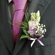 Resting against the deep, inky fabric of a tailored suit, this boutonnière feels like a small poem in flowers, crafted with the kind of care you might find in a trusted Brent Cross florist. At its centre, soft lilac and pale lavender blossoms open in layered petals, their colours somewhere between springtime heather and the fading sky over the North Circular at dusk. Tiny clusters of white baby's breath gather around them like a cloud of fine lace, each minuscule flower adding lightness and a touch of innocence. Curved, polished green leaves cup the composition, their surfaces smooth and reflective, shaping a gentle frame that keeps the blossoms nestled securely against the lapel. Slim, arching strands of vivid green grass rise from the base, giving the arrangement an airy, almost windswept feeling, as if a breeze from nearby Brent Cross Shopping Centre gardens had run through it moments before the photograph was taken. A sheer lavender ribbon binds the stems, its soft shimmer mirroring the tie tucked neatly beneath the jacket, creating an elegant conversation between fabric and flower. You can almost imagine a sweet, delicate fragrance lifting from the blooms-something fresh, like early morning walks along Hendon Way when the air is cool and full of promise. Designed for a wedding, prom, or special evening reception, the boutonnière feels both modest and full of emotion, an emblem of romance expressed on a miniature scale. Every line and curve reflects a florist's steady hand and thoughtful eye, turning a few simple stems into a wearable keepsake of the day.
