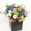 With its harmonious blend of colour, texture, and modern design, this premium floral arrangement from Brent Cross Flowers feels right at home in the heart of North London. The bouquet rises in a generous, rounded display from a sleek matte black square box, the dramatic container accentuating the riot of blossoms above. Towering hydrangeas in periwinkle and deep blue set a plush, cool-toned stage for lavish lavender and blush pink roses that softly unfurl at the centre, their layered petals offering an air of affection and serenity. Creamy lisianthus and golden-eustoma blooms introduce height and luminosity, their trumpet-shaped forms gleaming among the fresh lime green accent clusters and glossy foliage. Touches of feathery, baby pink astilbe flutter above, lending a whimsical movement reminiscent of botanical walks along the banks of the River Brent. Each element is carefully balanced and positioned, the arrangement illuminated by bright natural light that amplifies its dynamic hues against a pristine white background. Perfect for marking milestones, delivering comfort, or elevating local interiors from Staples Corner to Brent Cross Shopping Centre, this bouquet radiates both sophisticated charm and heartfelt celebration-an ideal floral gift for any season or occasion.