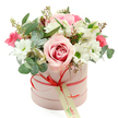 In the centre of the image, a charming floral hatbox arrangement is displayed against a spotless white background, giving it a sense of clarity and focus that mirrors a well-kept Brent Cross interior. The box itself is cylindrical and soft pink, with a smooth, matte surface that gently reflects the light. It is wrapped by a slim coral-red satin ribbon, neatly tied into a small bow at the front. A cream gift tag, decorated with green, handwritten-style lettering, dangles from the bow, hinting at a personal message or the careful touch of a local florist. The flowers rise above the box in a rounded dome, appearing abundant and carefully shaped. A prominent rose in pale pink, its centre deepening into a richer pink, sits at the heart of the composition, its velvety petals opening wide. Around it, pure white chrysanthemums or daisy-like blooms with bright green centres cluster closely, bringing freshness and a hint of playful charm. Threaded among them are white alstroemeria, their soft petals marked with fine speckles, and smaller deep pink flowers - possibly miniature roses or lisianthus - that add bright, romantic accents. The bouquet is finished with a generous selection of greenery: rounded silver-green eucalyptus leaves that arc gracefully, darker foliage that fills the gaps, and a few sprigs topped with tiny buds, giving an impression of natural growth. The result is a sophisticated yet approachable design, perfectly suited to same-day flower delivery around Brent Cross, from apartments near the shopping centre to family homes along Hendon Way, for occasions ranging from birthdays and anniversaries to thoughtful thank-you gestures or just-because surprises.