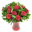 A lush, rounded bouquet of a dozen or more deep crimson roses fills the frame, each bloom opened fully to reveal tightly spiralled, velvety petals that graduate from a rich wine-dark centre to glossy outer edges. The scent seems almost audible in the image - a heady, classic rose perfume that pairs with the visual warmth of the petals. Dark green foliage is woven generously through and around the roses, small glossy leaves such as Ruscus or Salal forming a dense, verdant backdrop while broader, emerald Aspidistra-like leaves are artistically folded into elegant loops and spirals, their smooth, architectural curves lending a contemporary tension against the timeless softness of the rose heads. The flowers sit in a tall, translucent ruby-red glass vase whose subtle twisted or ribbed surface creates vertical lines that catch and reflect the even studio light, amplifying the bouquet's deep reds and adding a jewel-like gleam. The vase narrows at the waist and flares gently at the mouth, visually supporting the weight of the blooms while keeping the composition slender and refined. Set against a clean white background, the contrast is sharp and dramatic: the crimson roses and glossy greens appear almost three-dimensional, isolated and precious as a gift. The overall impression is romantic and sophisticated - an arrangement perfectly suited as an anniversary centrepiece, a Valentine's offering, or a luxurious personal gift. As a florist who works locally around Brent Cross and nearby Cricklewood, I'd imagine this bouquet arriving with the quiet confidence of careful handling and same-day freshness for a loved one in the neighbourhood.