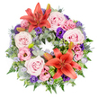 A beautifully composed circular funeral wreath rests against a clean white background, its design both soothing and visually rich. The ring is built on a generous bed of mixed greenery, combining rounded eucalyptus-like leaves, light and airy fern fronds, and deeper green foliage that together create a textured, natural frame. Two prominent coral-orange Asiatic lilies act as the main focal points, placed on opposing sides of the wreath so that their wide, star-shaped petals and prominent stamens stand out warmly against the greens. Around these lilies, groups of soft blush and lavender-pink roses are clustered, their velvety petals unfolding in elegant spirals that give the arrangement a tender, comforting feel. Threaded throughout the roses, ruffled lisianthus blooms in pastel pink and deep, royal purple add depth and subtle variation, their cup-shaped flowers lending a gentle sense of movement. Rising from the softer textures, small spiky blue-grey eryngium heads introduce a cool accent and a hint of structure, while fine sprays of delicate lavender-blue filler flowers float between the larger blooms like a soft mist, helping to blend the colours into a harmonious whole. Hidden among the greenery, a few unopened lily buds offer a quiet suggestion of new life, even as the wreath honours a life that has passed. Near one of the lilies, bright green leaves are folded and curved in a sculptural way, showcasing skilled floristry you might expect from a dedicated Brent Cross florist. The overall impression is one of calm respect and gentle beauty, an arrangement perfectly suited to a memorial service or funeral in Brent Cross or for delivery to families across the surrounding North London areas.