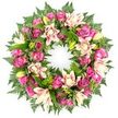 A sumptuous circular sympathy wreath of roses and lilies is pictured against a bright, uncluttered white background, drawing attention to every petal, stem, and leaf. The wreath is crafted as a complete ring, richly filled so that flowers gently overlap, creating a seamless band of colour. Central to the design are roses in varying shades of pink, from deep magenta and bold fuchsia to soft, pastel blush. Their petals are thick and velvety, some blooms wide open in full display, others half-open or still tightly formed buds, giving the arrangement an organic, evolving feel. Nestled among these roses are large double lilies, whose generous petals undulate outward. These lilies blend creamy whites with pale pinks and touches of peach, with subtle hints of darker pink along some edges, providing a painterly depth. Tucked between the open lilies are several bright yellow-green buds, upright and slender, suggesting the promise of more blooms to come. All the flowers are supported by a robust base of lush greenery: fine fern fronds extend in feathery arcs, while glossy deep green leaves fill the inner circle, adding weight and structure. The contrast between the dark greens, warm pinks, and the neutral white background makes the wreath appear vivid and three-dimensional, as if it were resting on a stand at a chapel or crematorium near Brent Cross. The overall effect is both dignified and gentle, a condolence wreath that feels personal and considered, well suited to honouring a life at funerals, memorial services, or celebrations of life across Brent Cross and the neighbouring North West London communities.