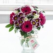 A vibrant mixed bouquet of fresh flowers stands proudly in a squat, clear, textured glass vase, set against a softly glowing white background that feels like daylight filtering through a Brent Cross apartment window. The arrangement is dominated by large gerbera daisies in intense fuchsia and magenta, their velvety petals circling deep, dark centres that draw the eye immediately. Around them cluster bicolour chrysanthemums, each petal tipped in rich purple before fading gently to white, framing bright yellow centres that add a cheerful sparkle. Nestled between these bold blooms are creamy white spray roses, some fully open and others still in tight bud, giving the arrangement a sense of movement and unfolding promise. Fine sprigs of white gypsophila, or baby's breath, create a soft, cloud-like layer, lightening the deeper tones and adding a delicate, airy texture. Lush, glossy green foliage peeks from the base and between the flowers, grounding the design and making the colours appear even more vivid. The vase is cinched at the neck with a rustic bow of natural twine, from which hangs a small white gift tag printed with a stylised pink rose icon and flowing script, hinting at a handwritten message for a birthday, thank you, or just-because surprise. The bright, even light highlights the crisp freshness of each petal and leaf, suggesting a bouquet freshly delivered to a home near Brent Cross Shopping Centre, ready to brighten a kitchen table or living room. The whole scene feels joyful yet refined, like a thoughtful floral gift chosen to lift someone's spirits on an ordinary North London day.